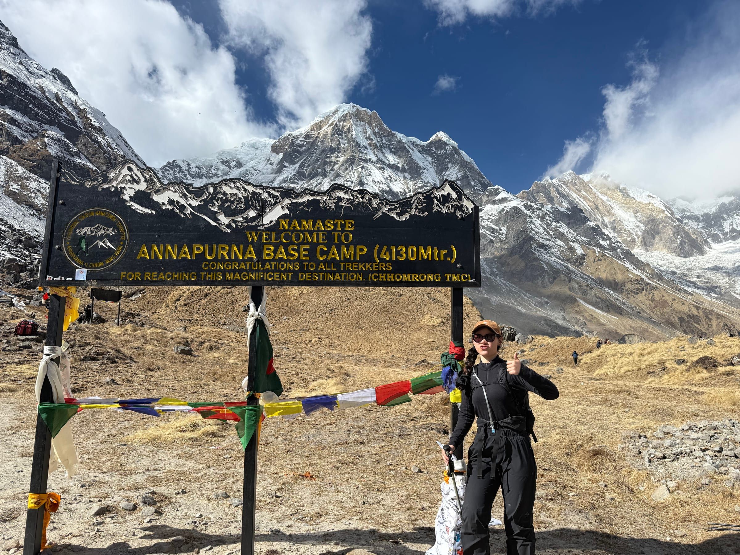 Standing beside the Annapurna Base Camp sign at 4,130m, snow peaks and prayer flags behind