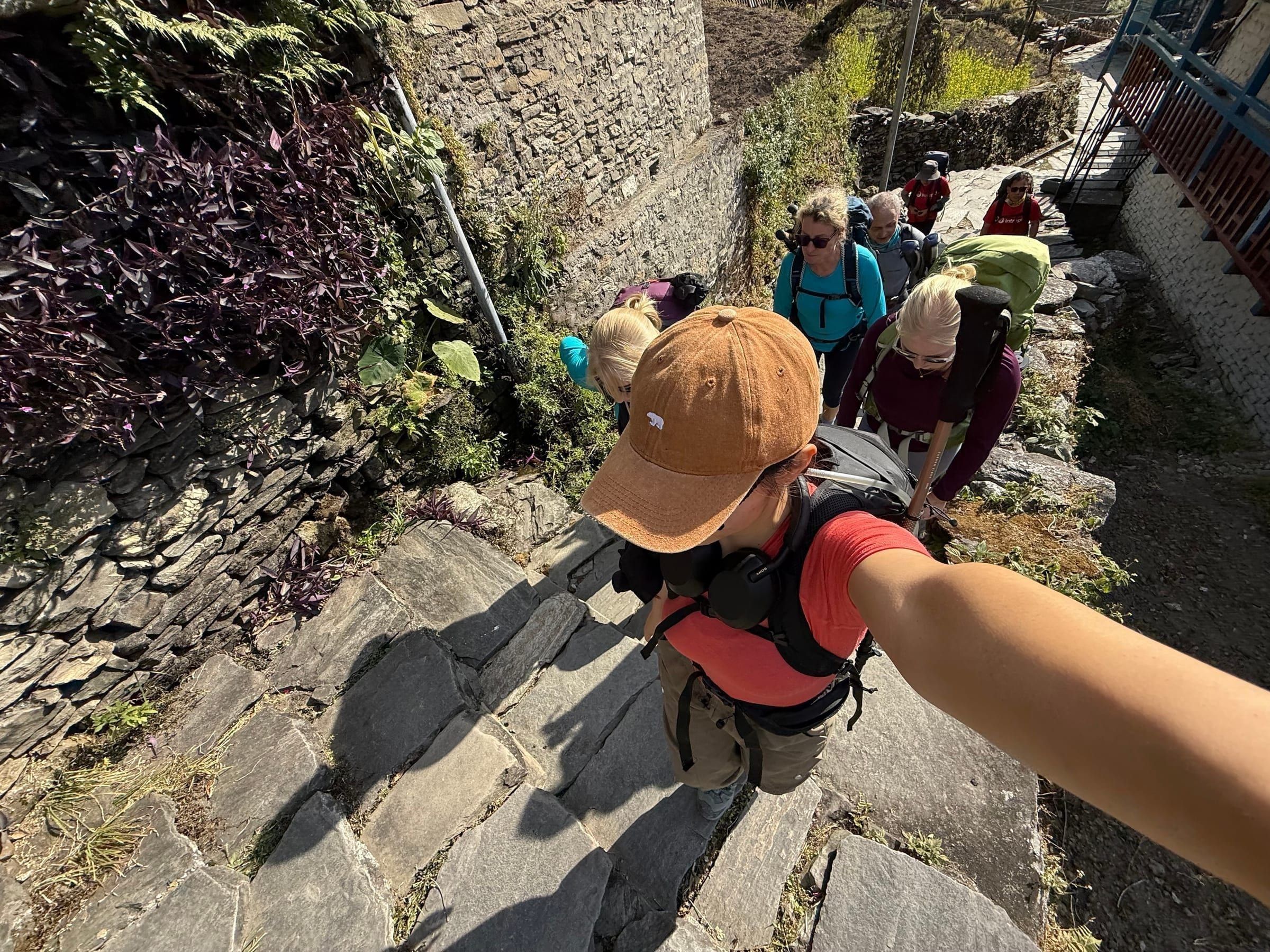 A trekker's selfie looking back at a group climbing stone steps through a village, backpacks and hiking poles in frame