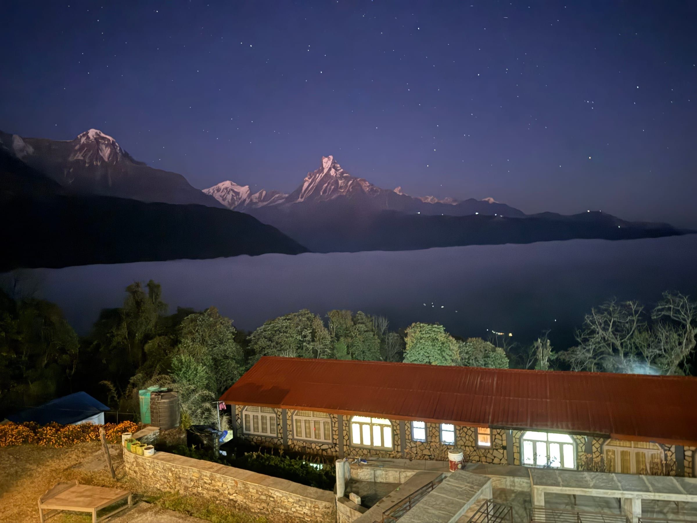 Full night sky thick with stars over a snow-capped peak, a lit teahouse building in the foreground