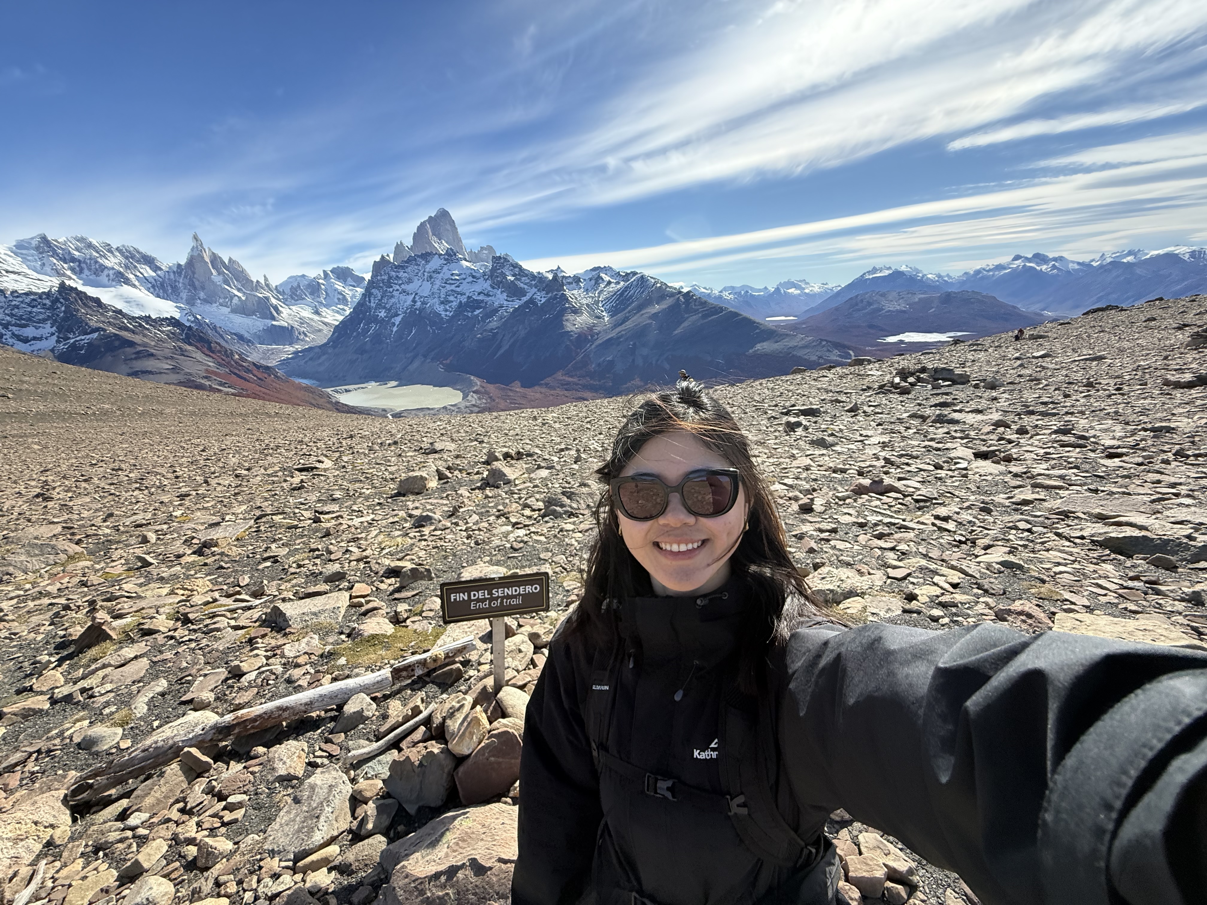 A woman in hiking gear smiling at the camera on a rocky trail, with the Fitz Roy and Cerro Torre massif rising behind her under a blue sky, a wooden "Fin del Sendero / End of trail" sign beside her