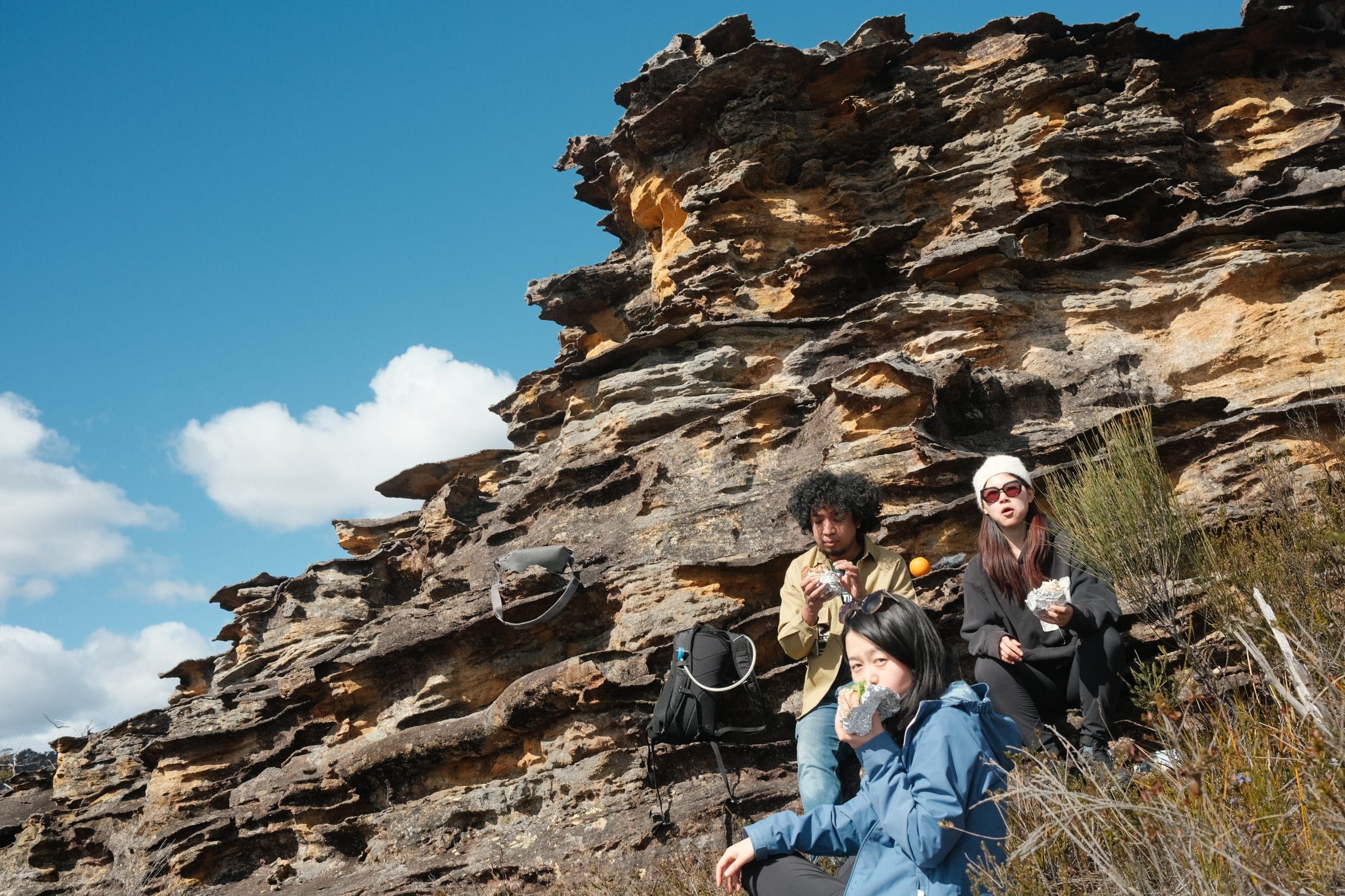Three friends sitting on a rocky sandstone ledge, eating foil-wrapped sandwiches with a cliff face behind them