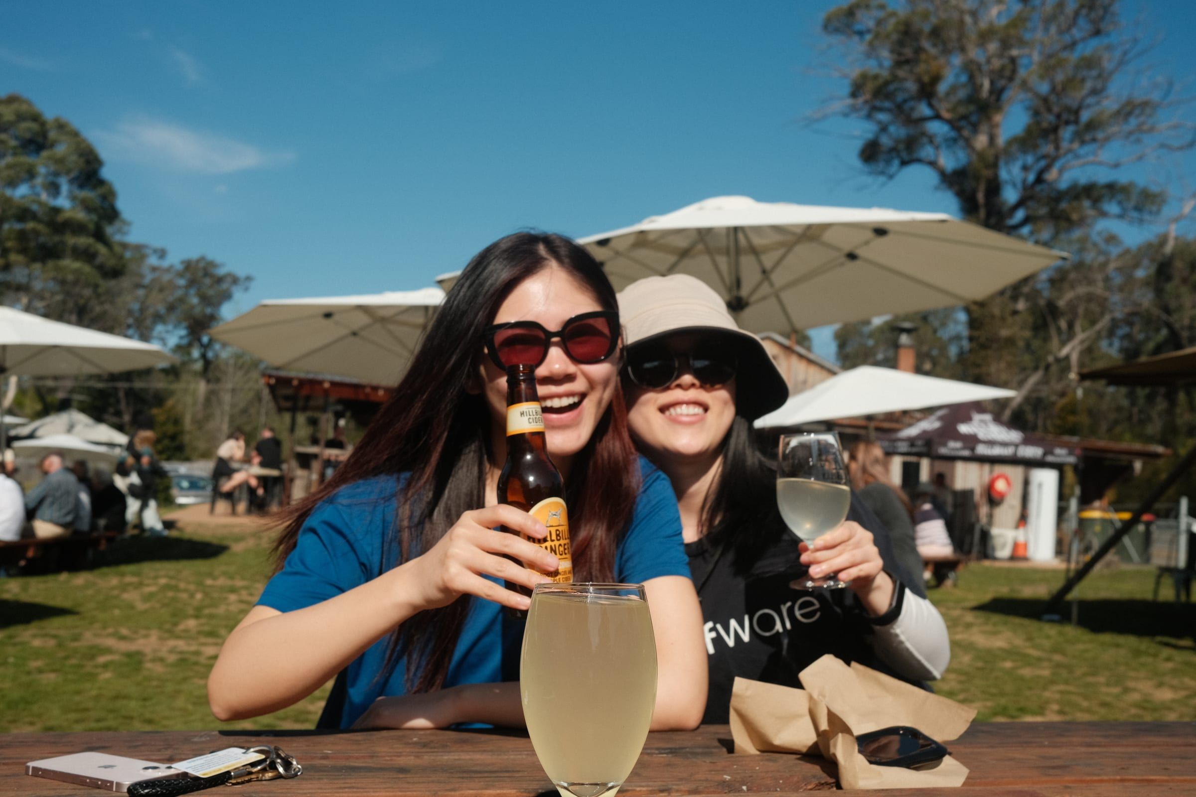 Two friends laughing over cider and wine at an outdoor table, white umbrellas and a cidery building in the background