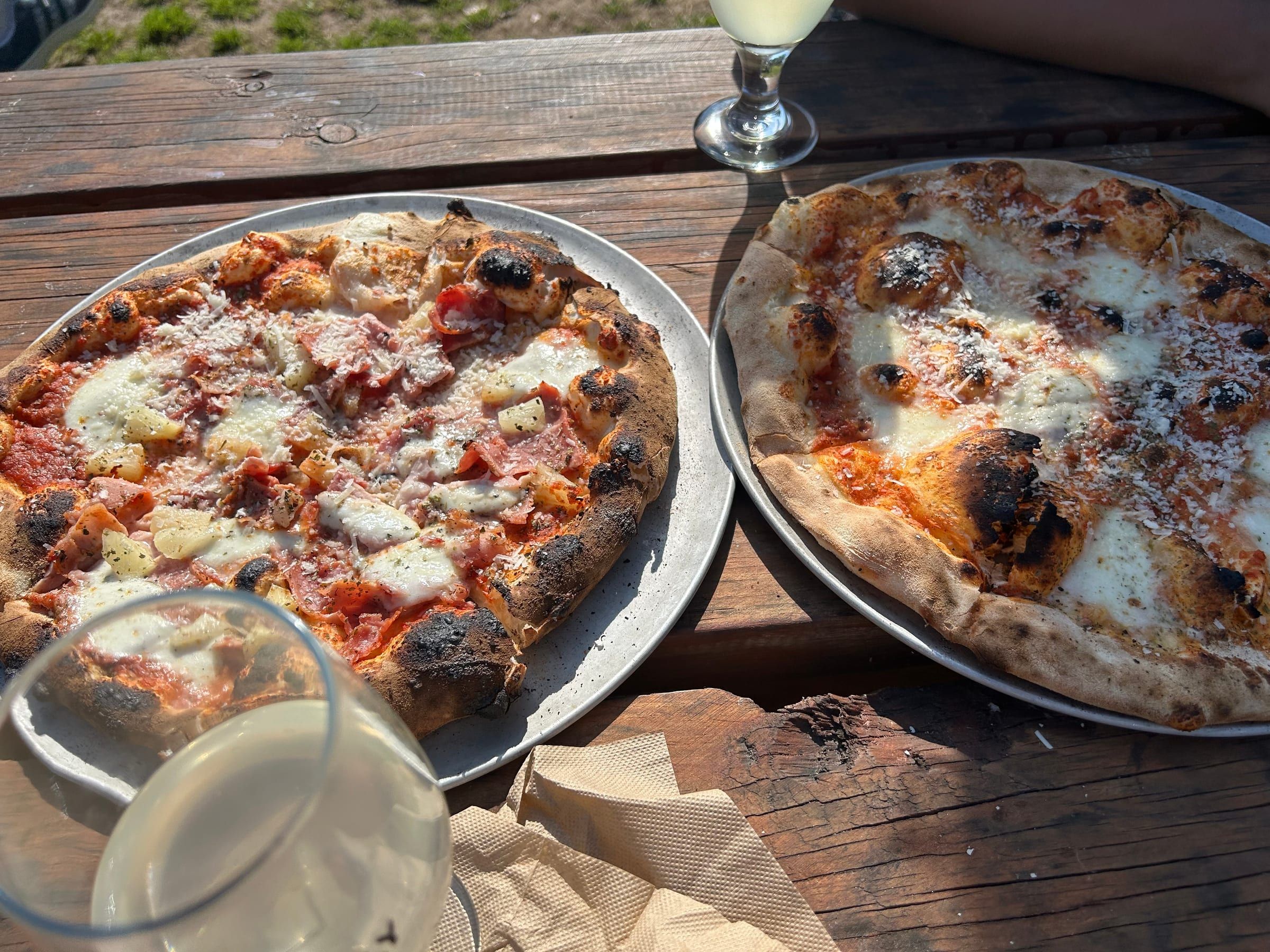 Two wood-fired pizzas on metal trays, glasses of cider and wine on a rustic wooden table