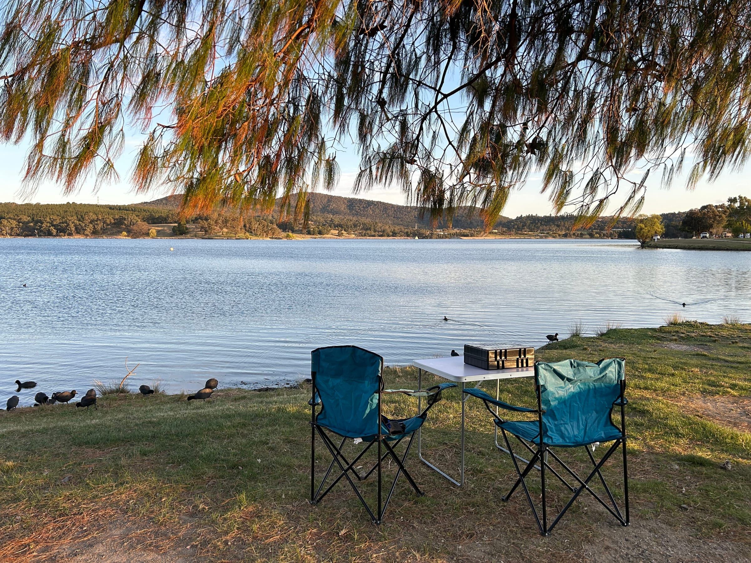 Two camp chairs and a small table set up on the grass beside a lake at dusk, ducks foraging nearby