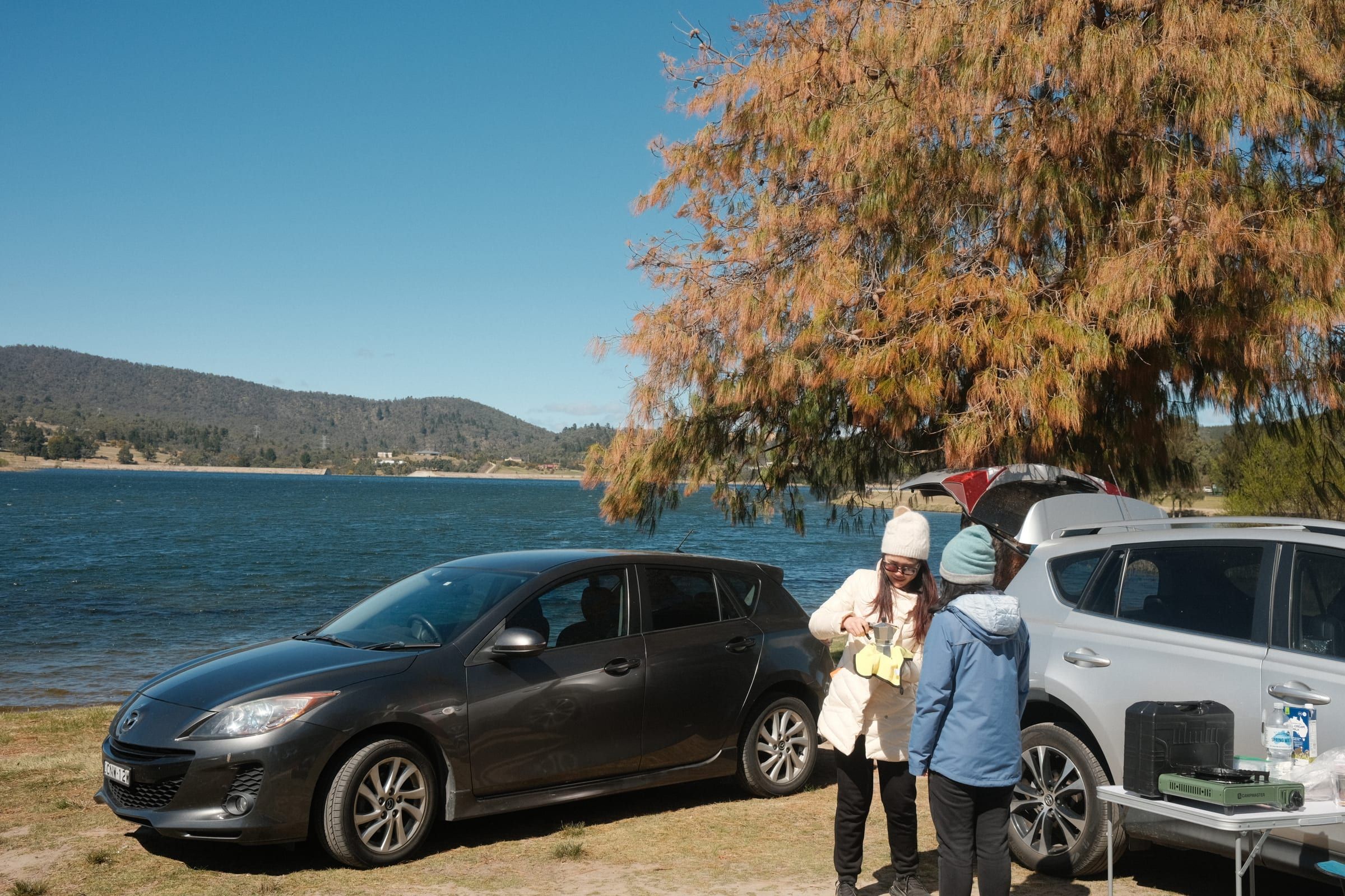Two friends making coffee with a stovetop moka pot beside parked cars at the lake edge