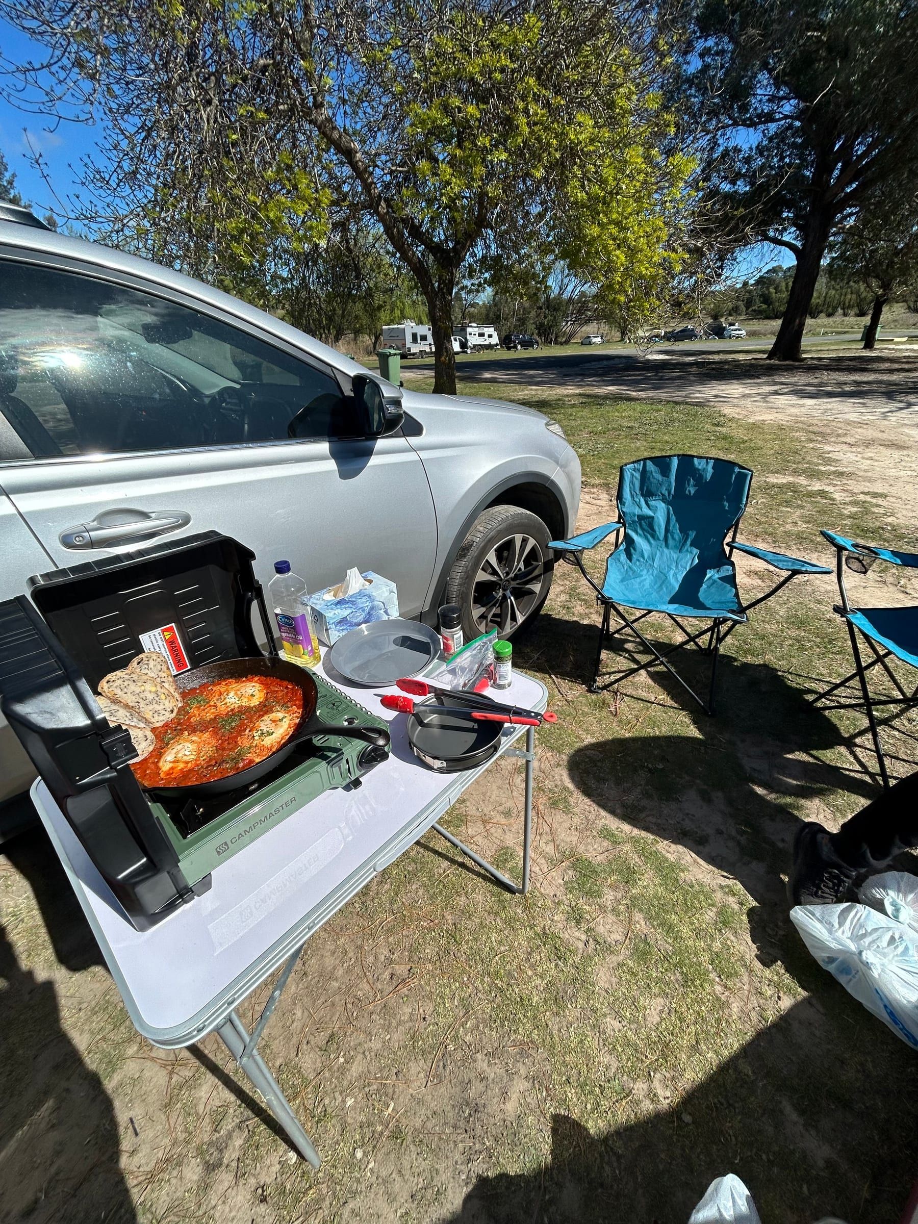 A cast iron pan of shakshuka with sliced sourdough, cooking on a portable stove at a camp table beside a car