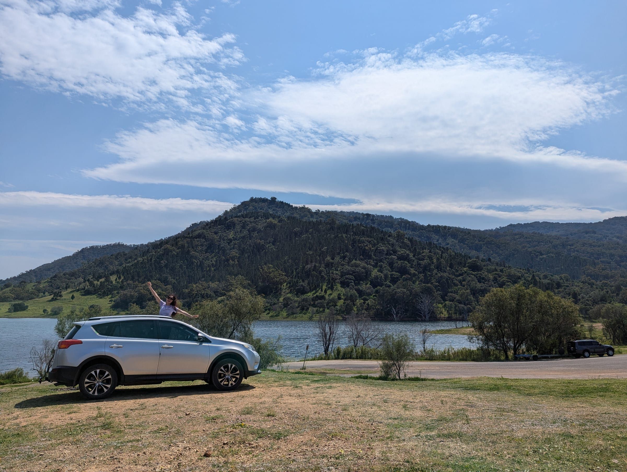 A silver SUV parked beside a wide lake, hills rolling into the distance under a clear blue sky