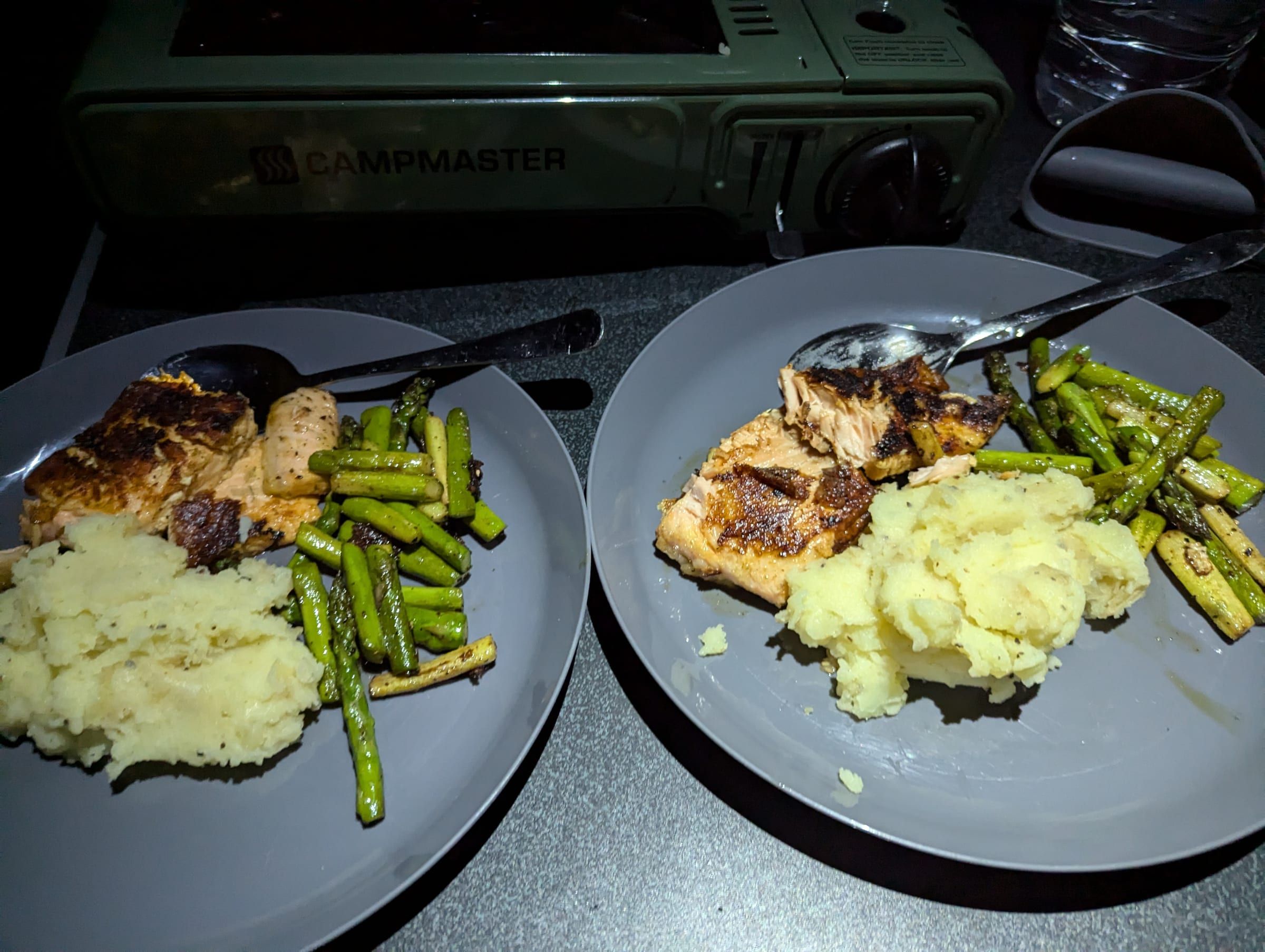 Two plates of seared salmon, mashed potato, and roasted asparagus, cooked on a portable camp stove