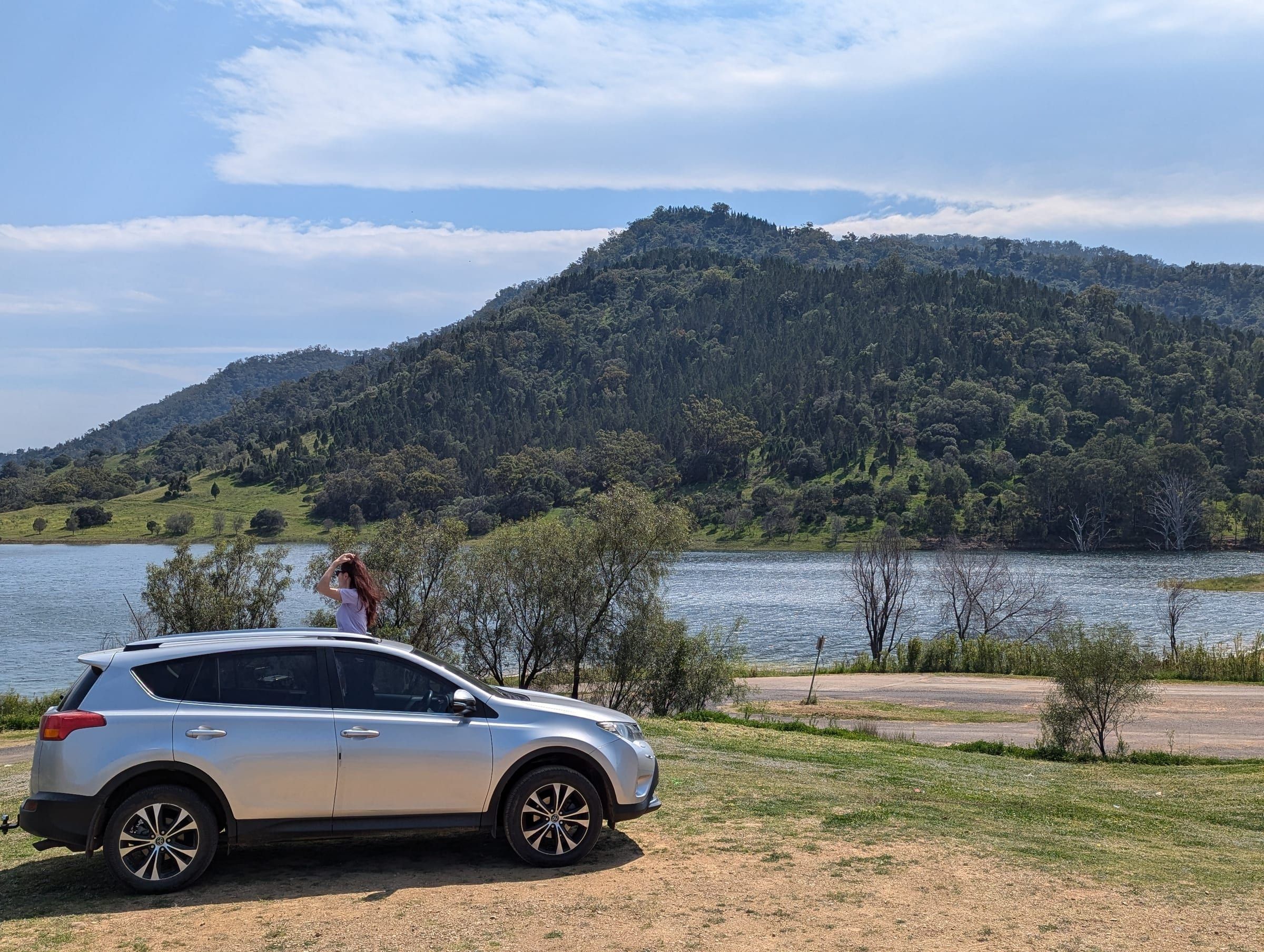 A person standing through a car's open sunroof, arms raised, overlooking a lake and green hills