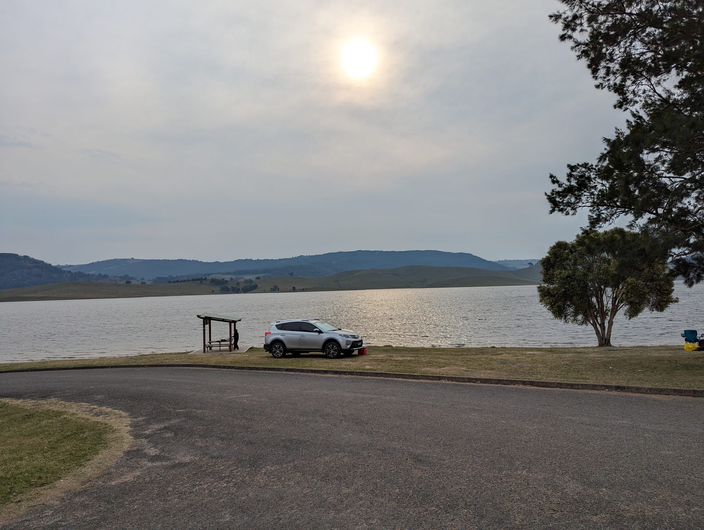 A car parked beside a wide lake in golden late-afternoon light, a large tree standing beside the water
