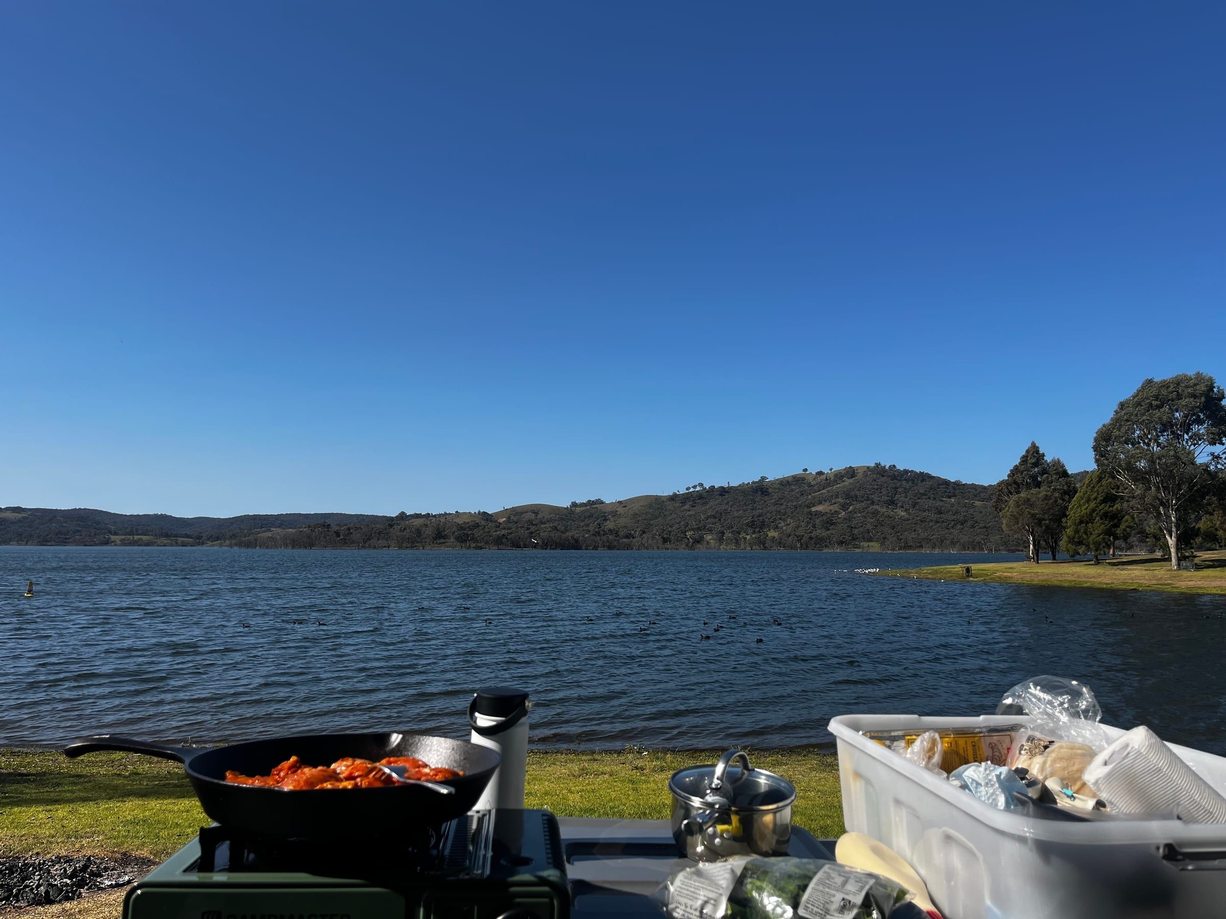 A cast iron pan of tomato-based stew cooking on a portable camp stove, with a wide lake and hills visible behind