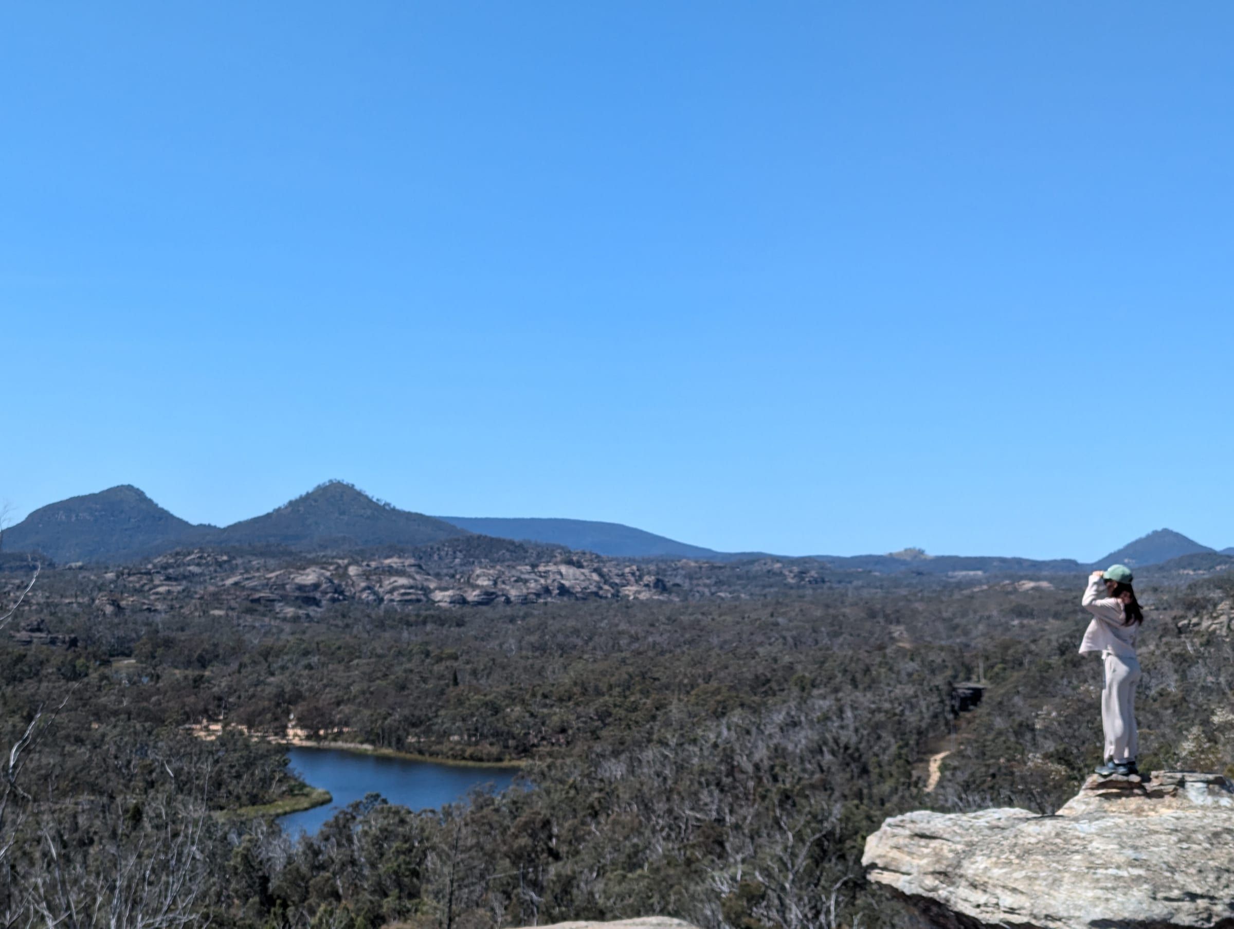 A person standing on a large rock outcrop, looking out over a forested valley with a lake visible below