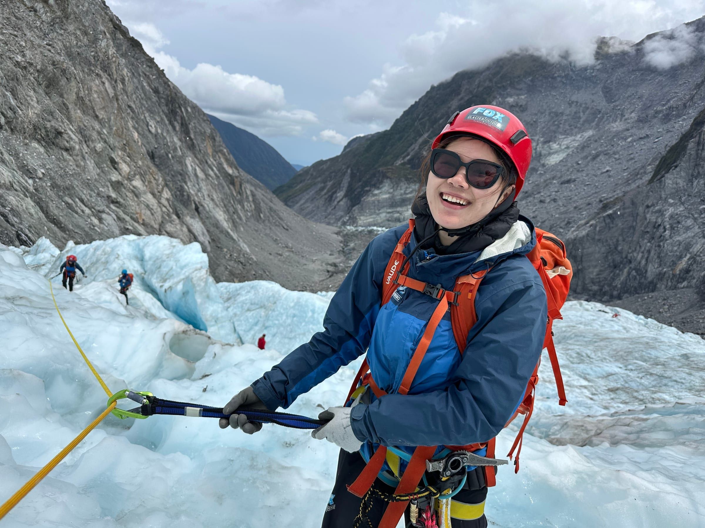 Roped up on the ice, wearing a helmet and harness, laughing mid-climb with the glacier valley behind