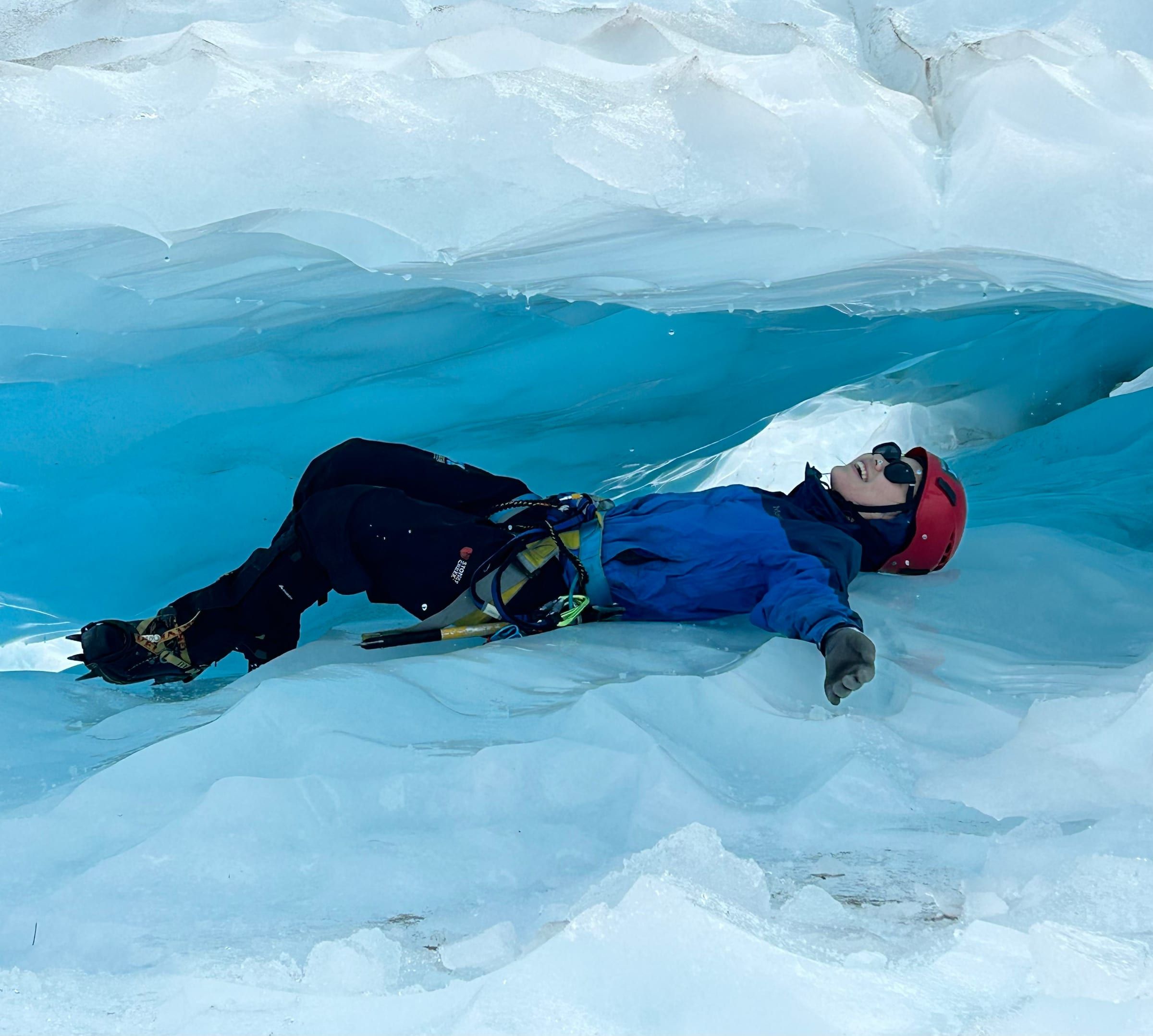 Lying inside a bright blue ice cave on the glacier, crampons visible, laughing up at the ceiling of ice