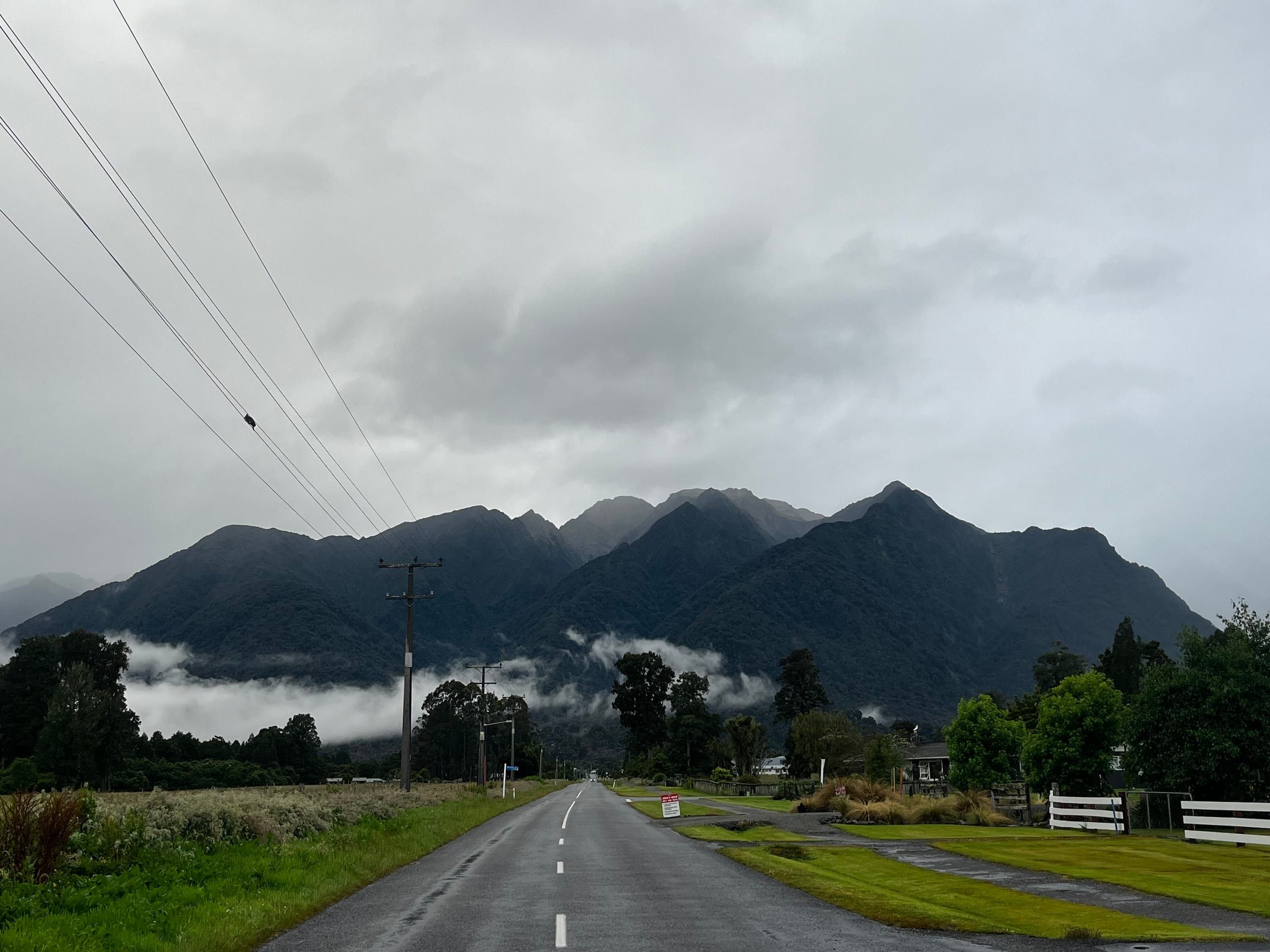 A quiet rural road leading toward dark, cloud-wrapped mountains under heavy evening light