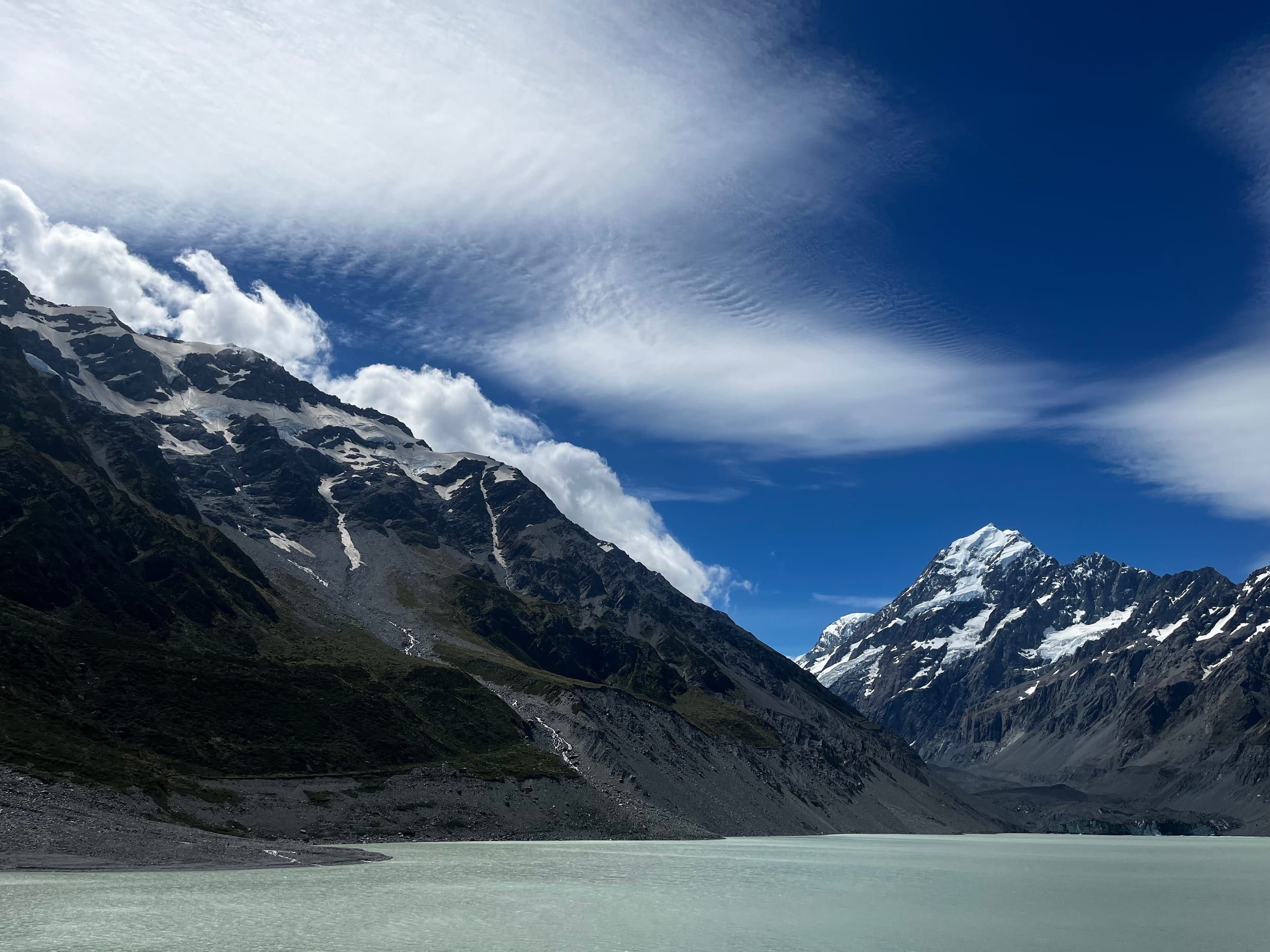A milky turquoise glacial lake at the base of Aoraki/Mt Cook, dramatic clouds streaming off the summit