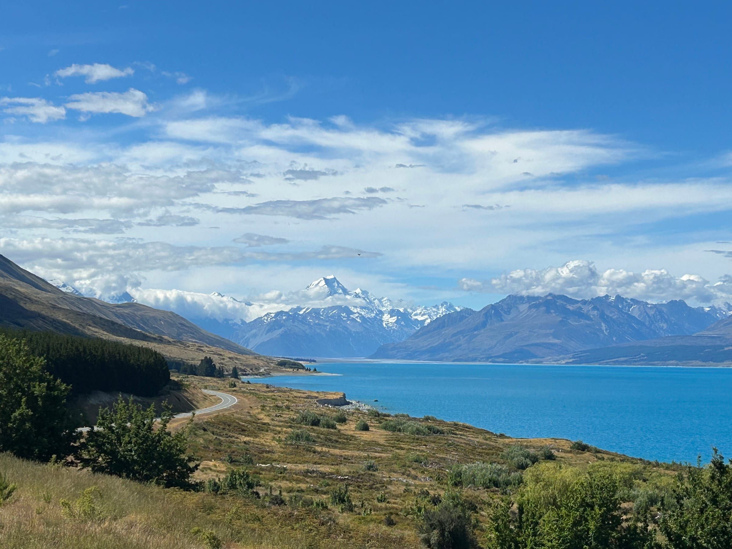Lake Pukaki's turquoise water stretching toward Aoraki/Mt Cook, a road curving along the shoreline