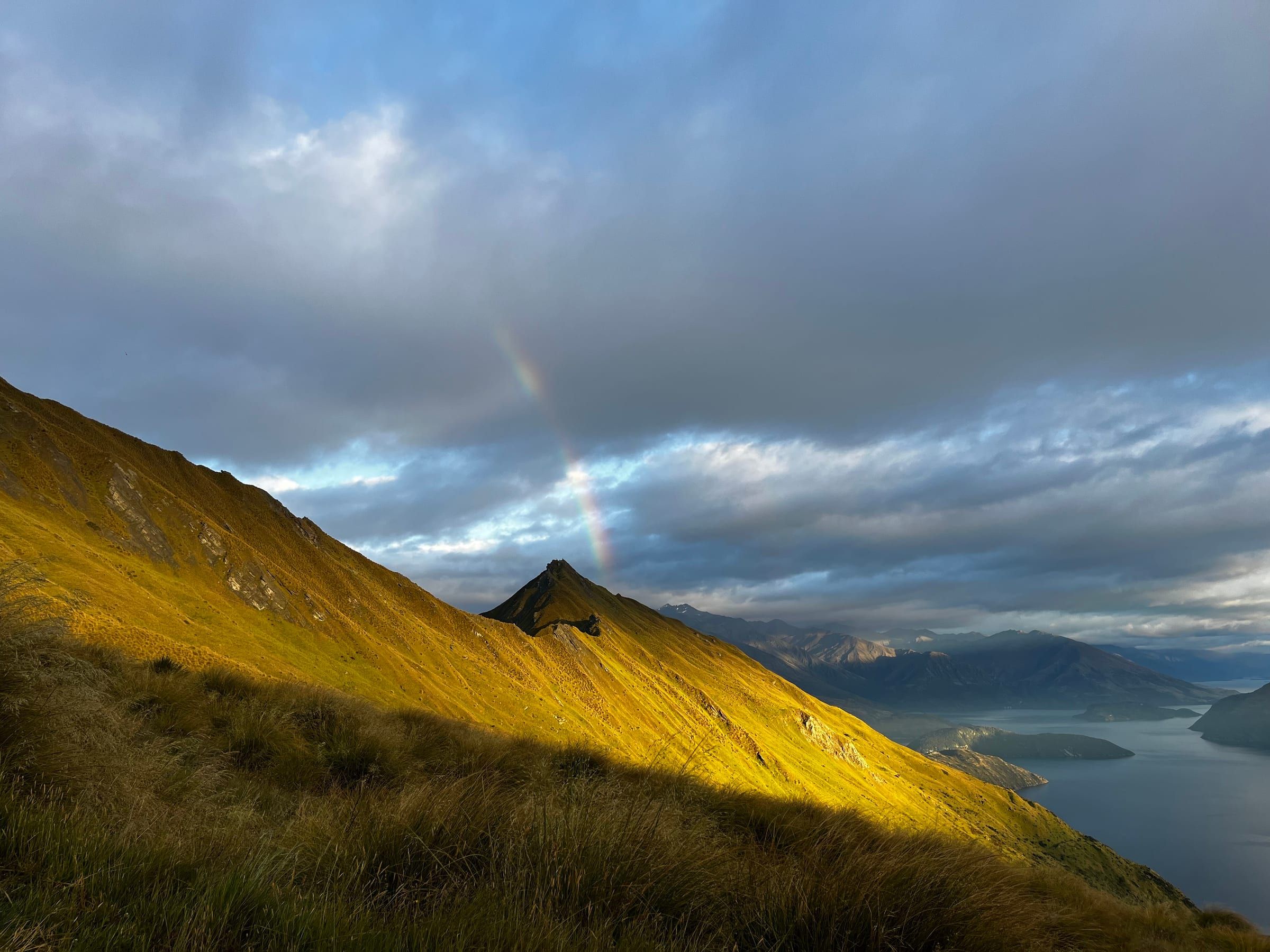 A rainbow arcing down toward a golden, sunlit ridge, lake and distant mountains below
