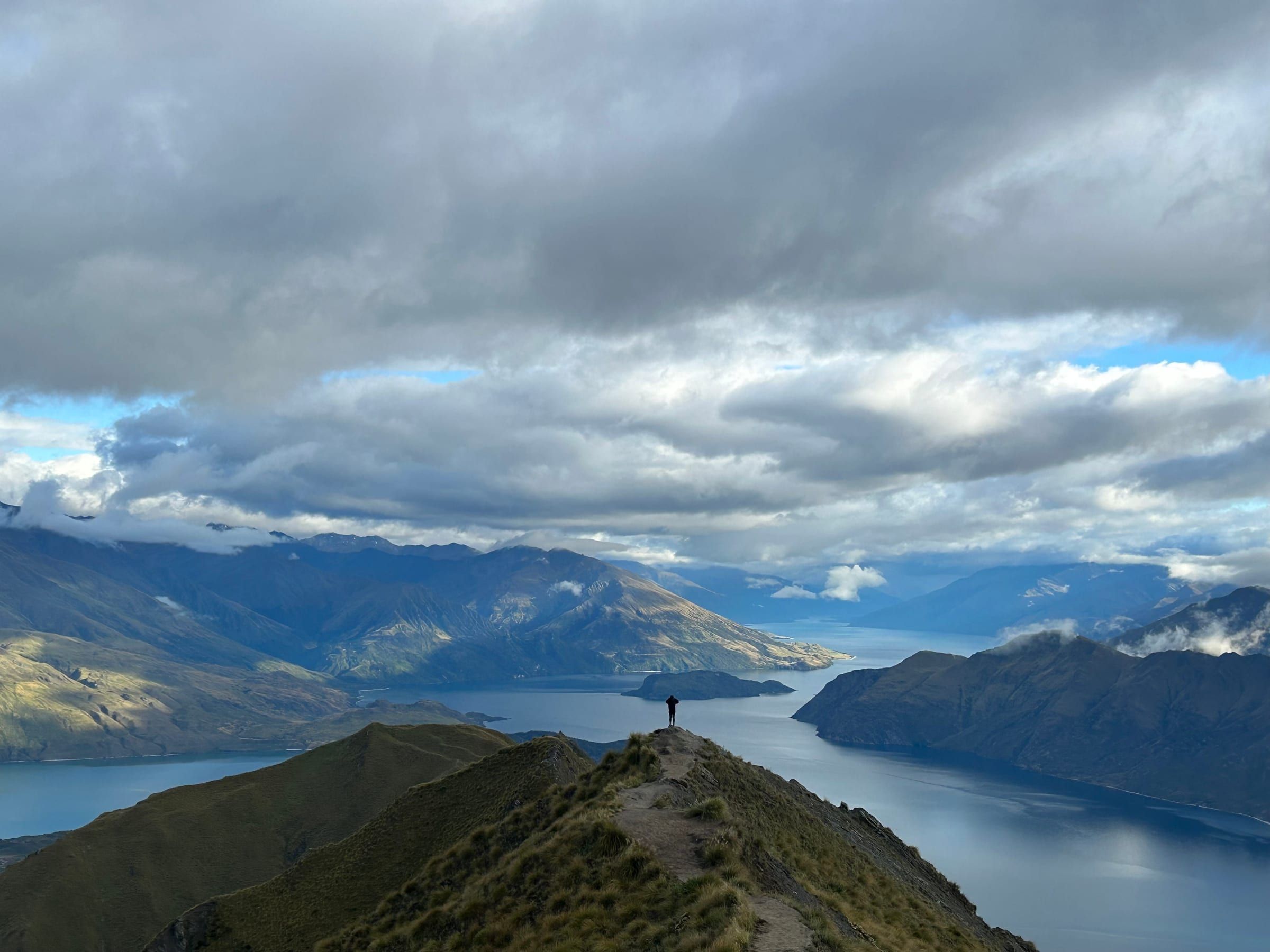 A lone figure standing silhouetted on the narrow summit ridge of Roy's Peak, Lake Wanaka stretching out below between mountain ranges