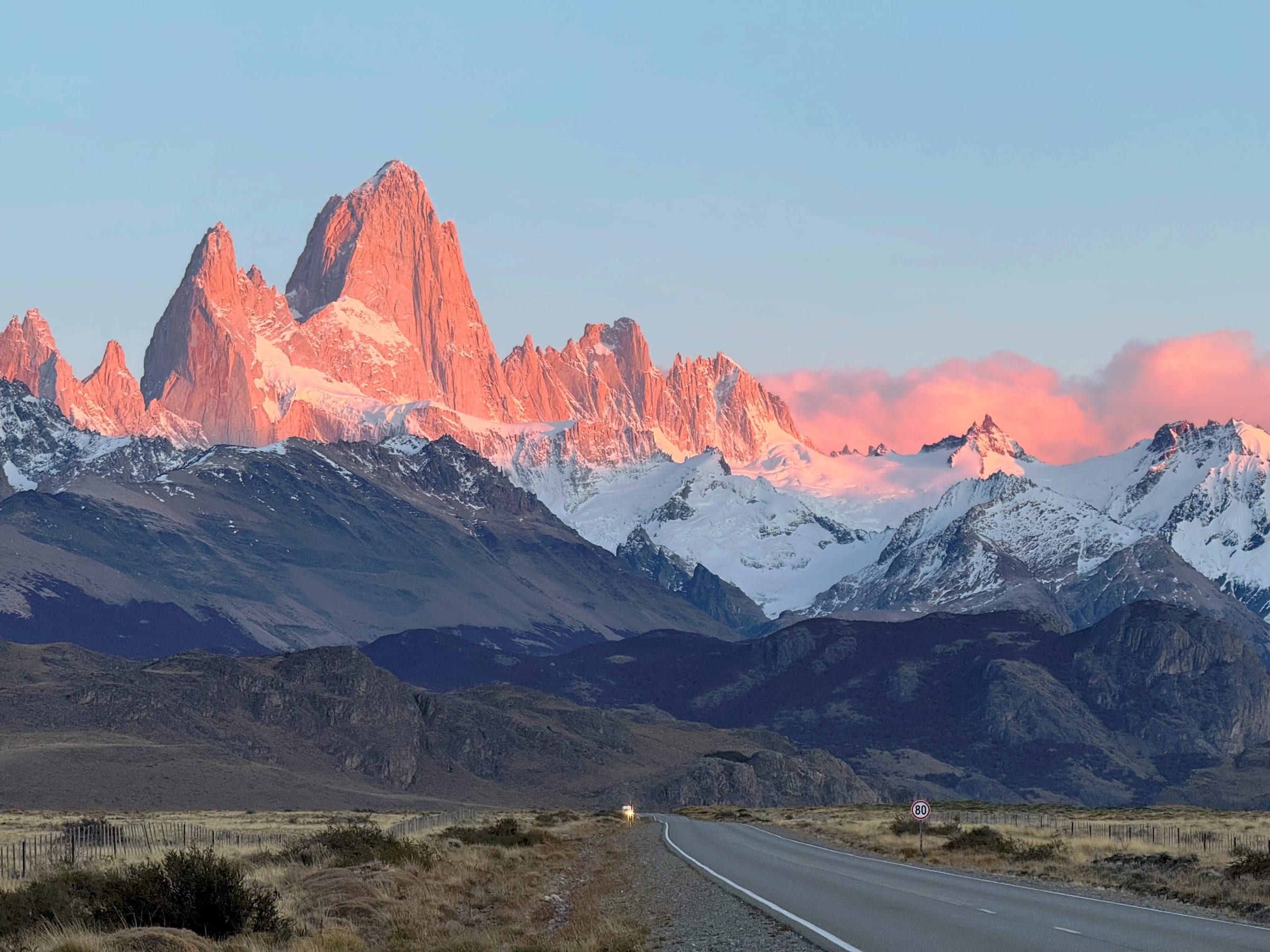 Fitz Roy at sunrise, granite spires glowing pink and orange above snowfields, empty road with speed sign in foreground