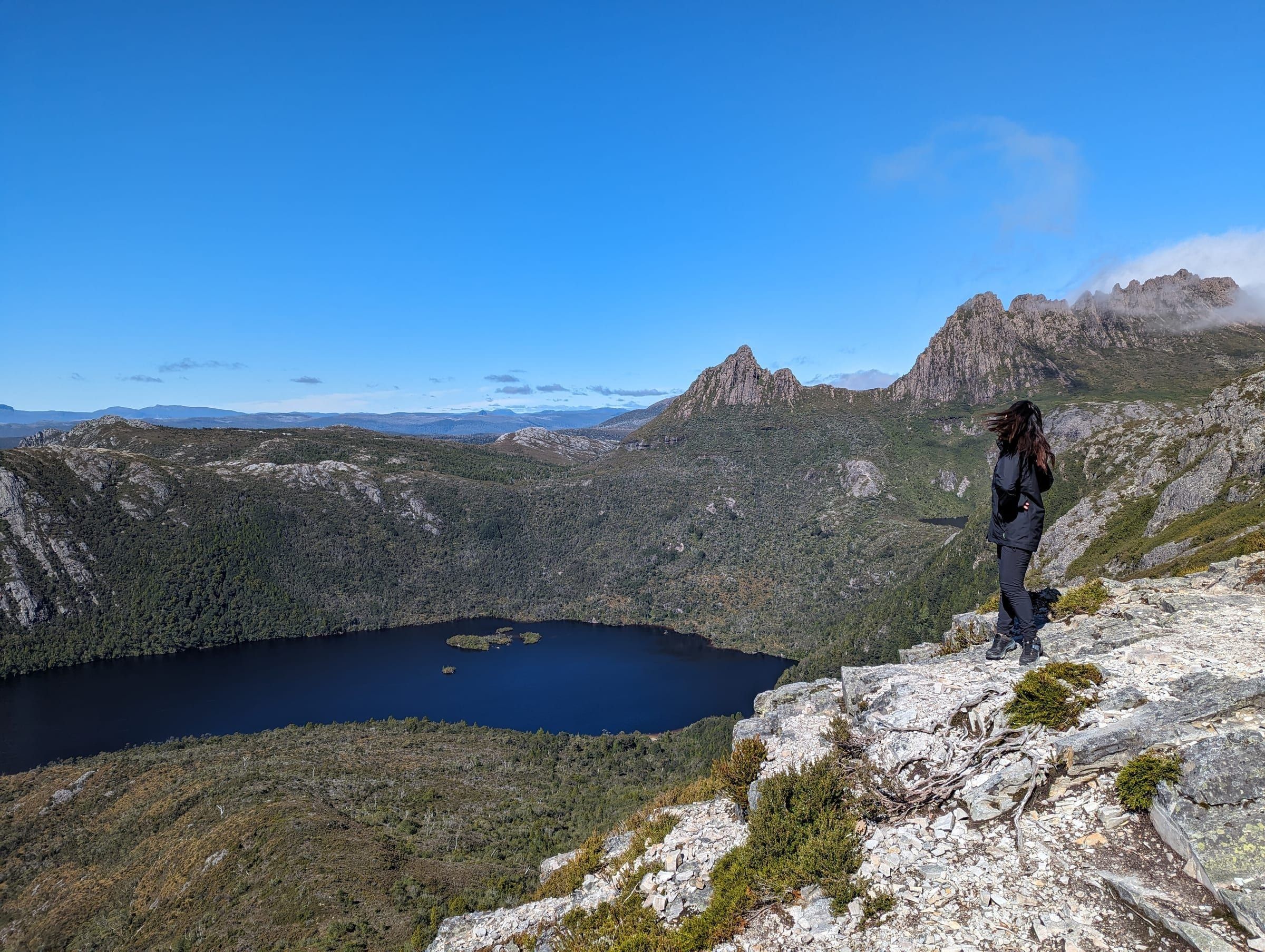 A hiker standing at the edge of a rocky lookout, Cradle Mountain's jagged peaks and a deep blue lake below