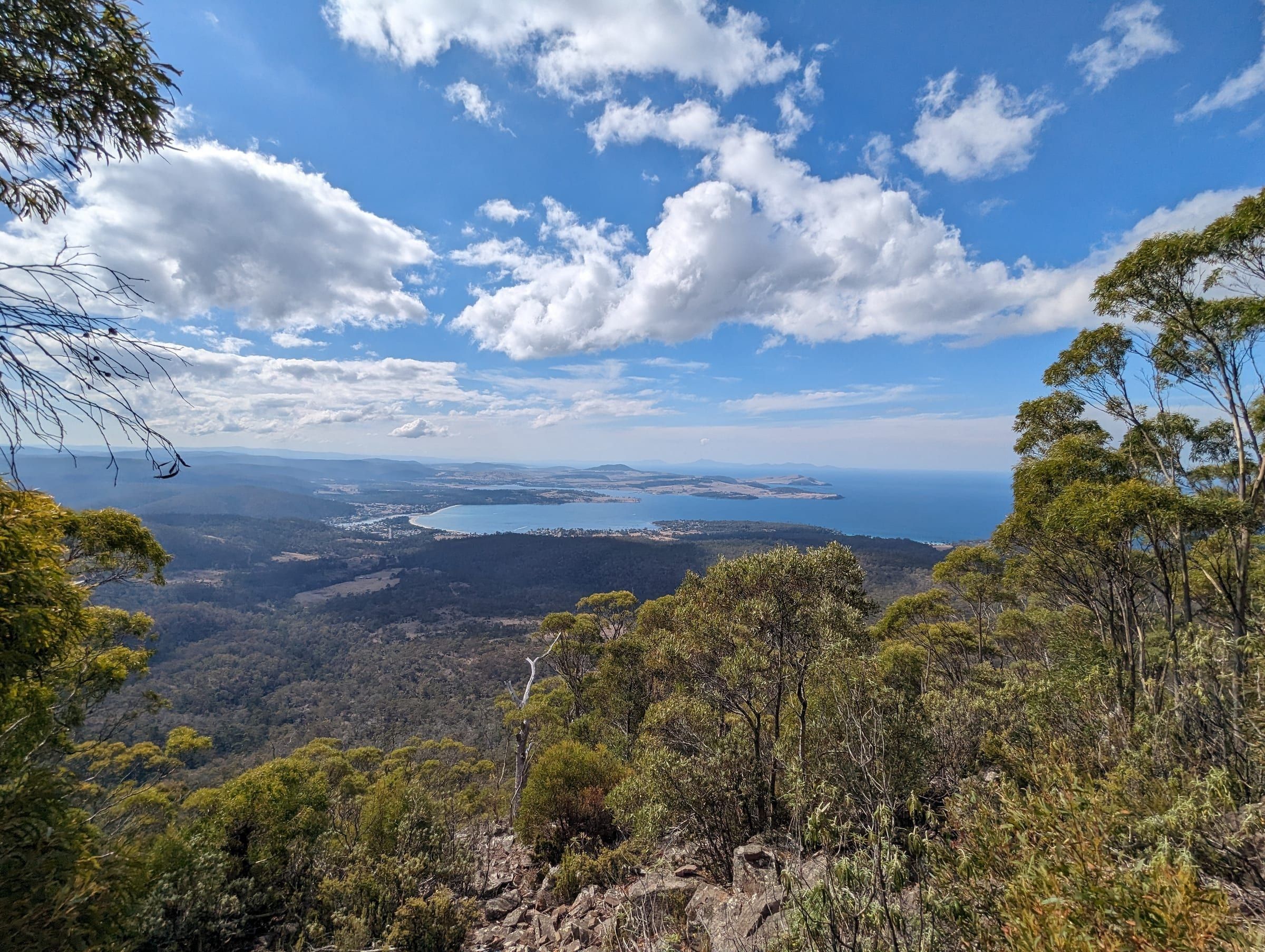 A sweeping view of a coastal bay and township from a high mountain lookout, clouds overhead