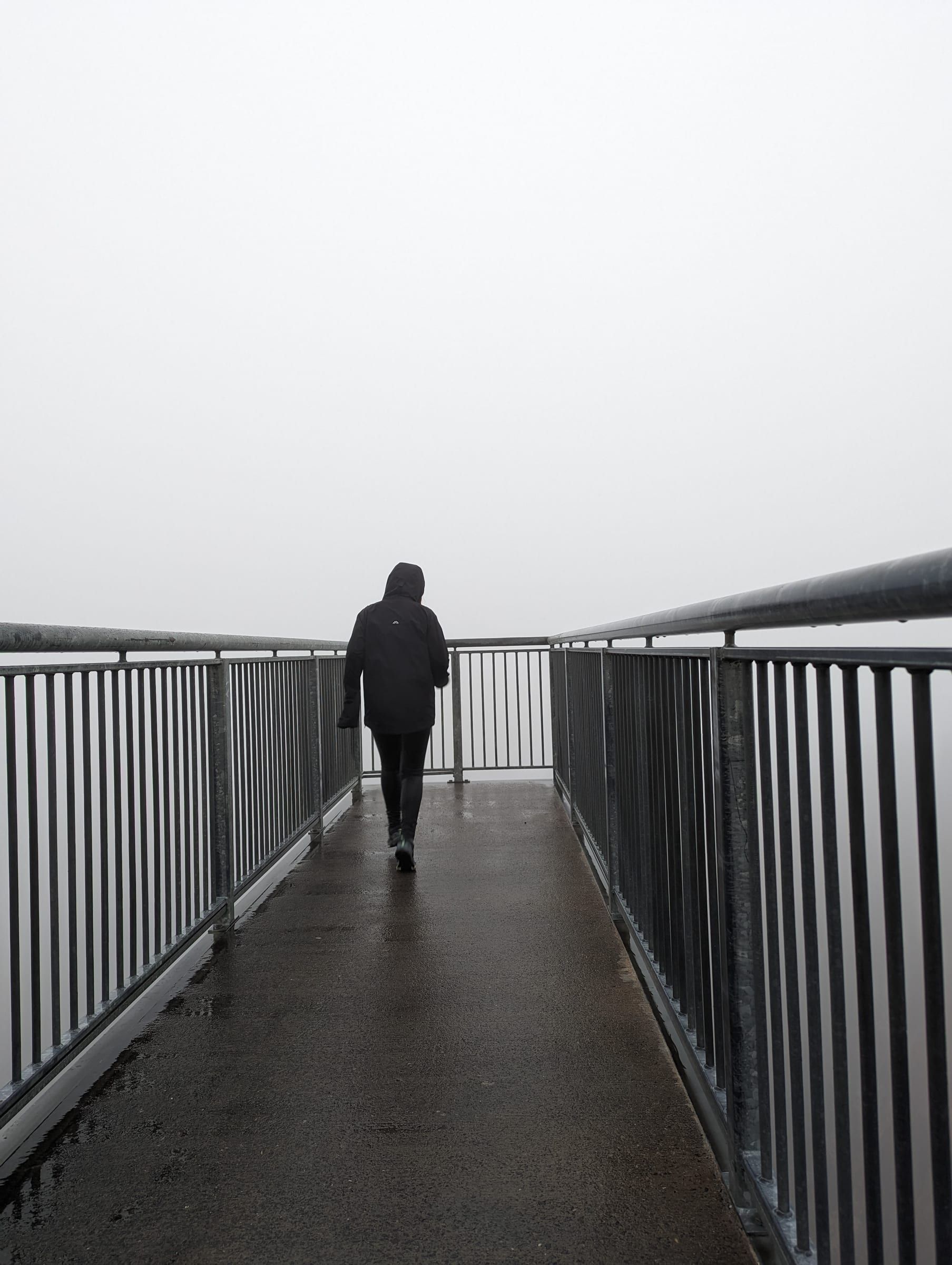 A figure in a rain jacket walking along a foggy viewing platform, visibility reduced to almost nothing