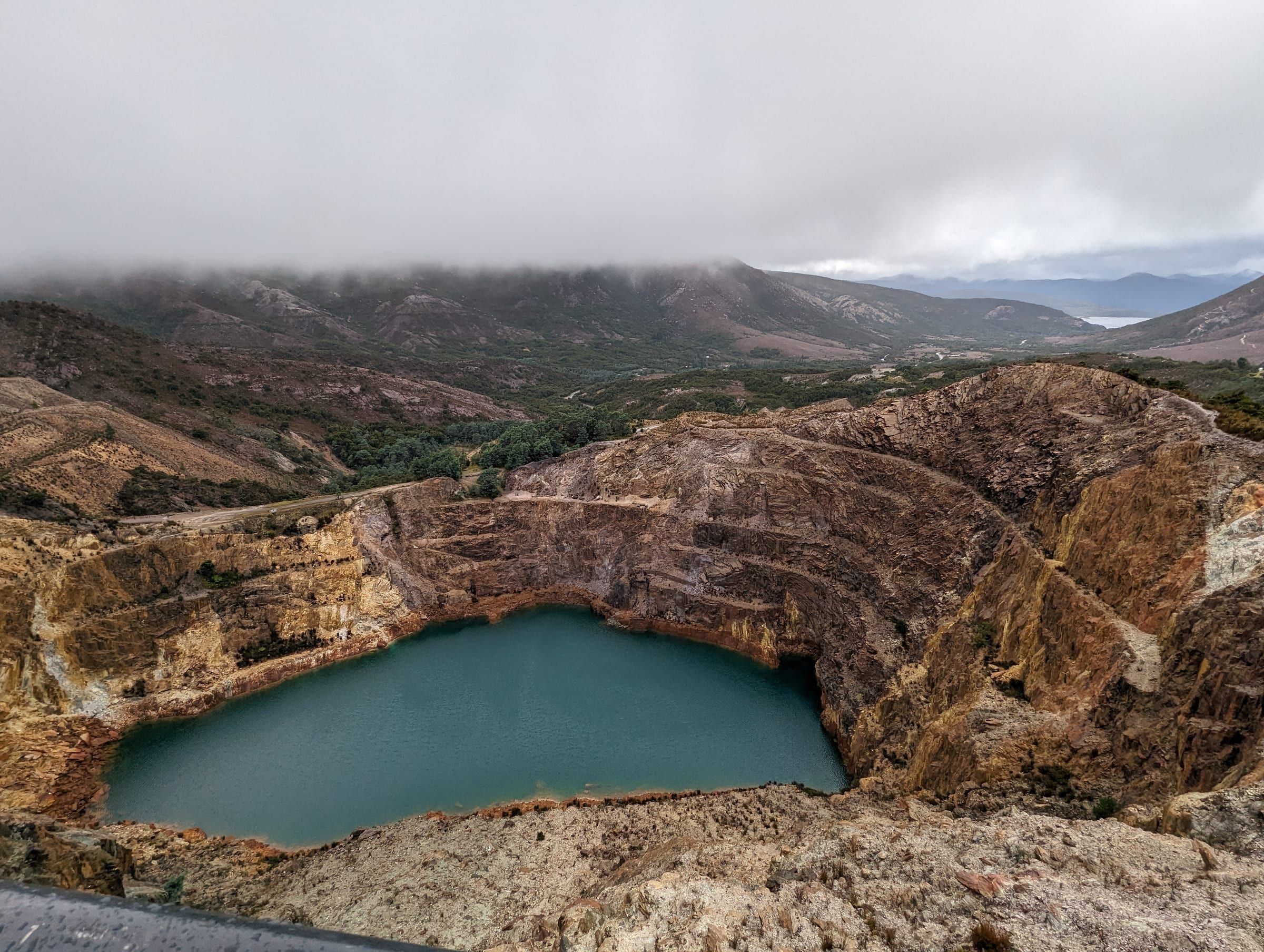 A vivid turquoise lake filling an old open-cut mine pit, fog rolling over the ridgeline above