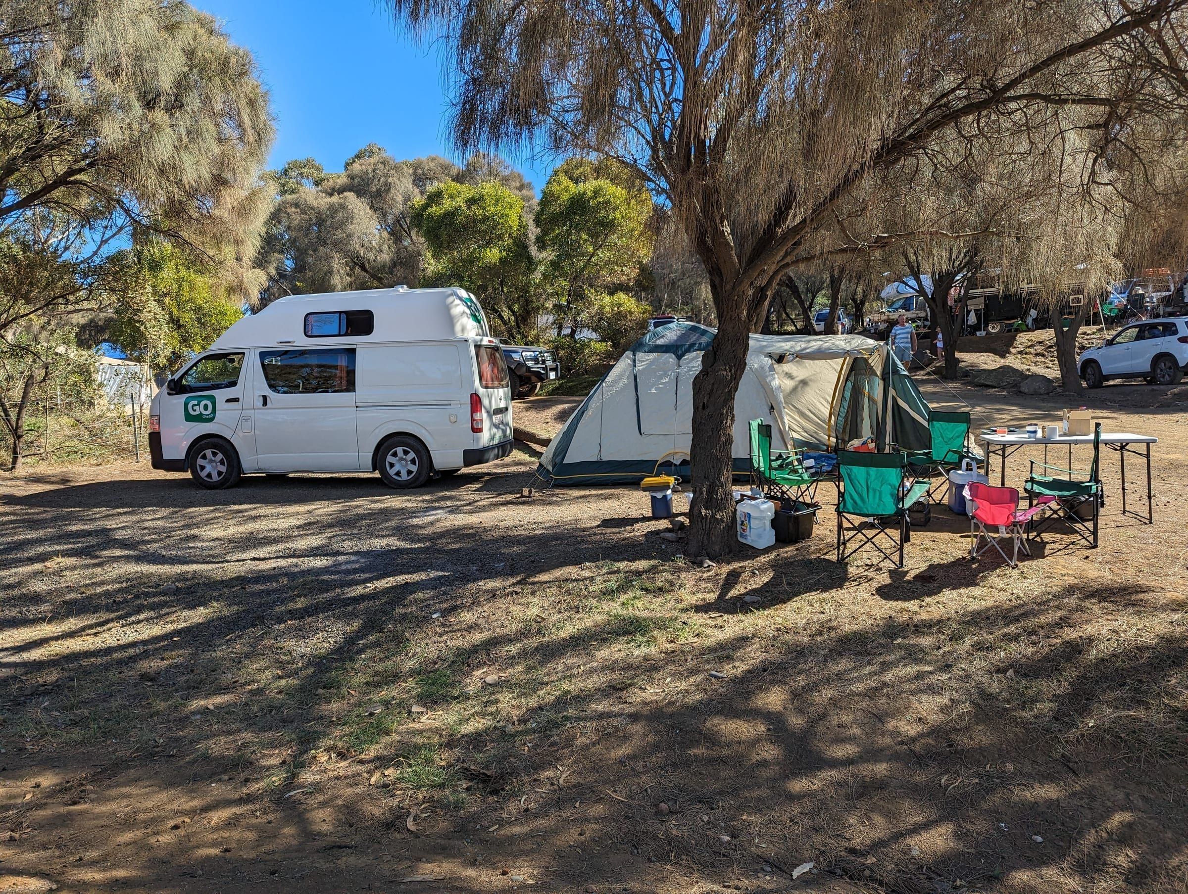 A campground packed with vans, tents, and camp chairs under shady trees