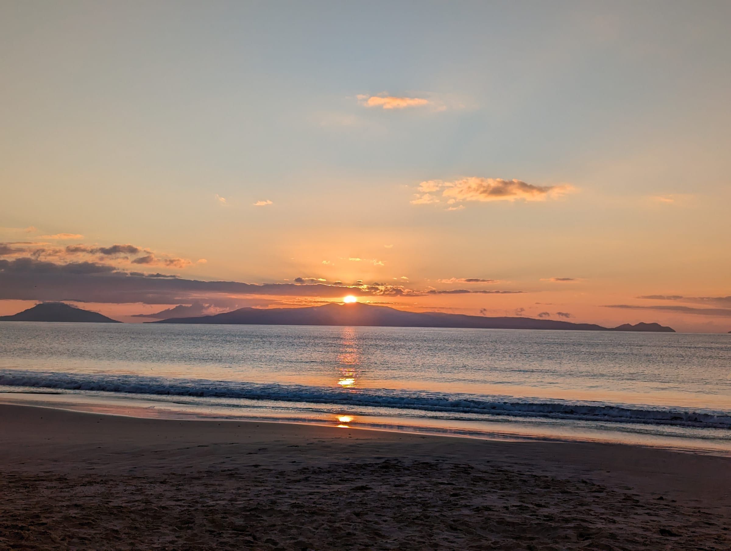 A wide sunrise over the ocean, the sun fully risen above distant mountains, waves rolling in