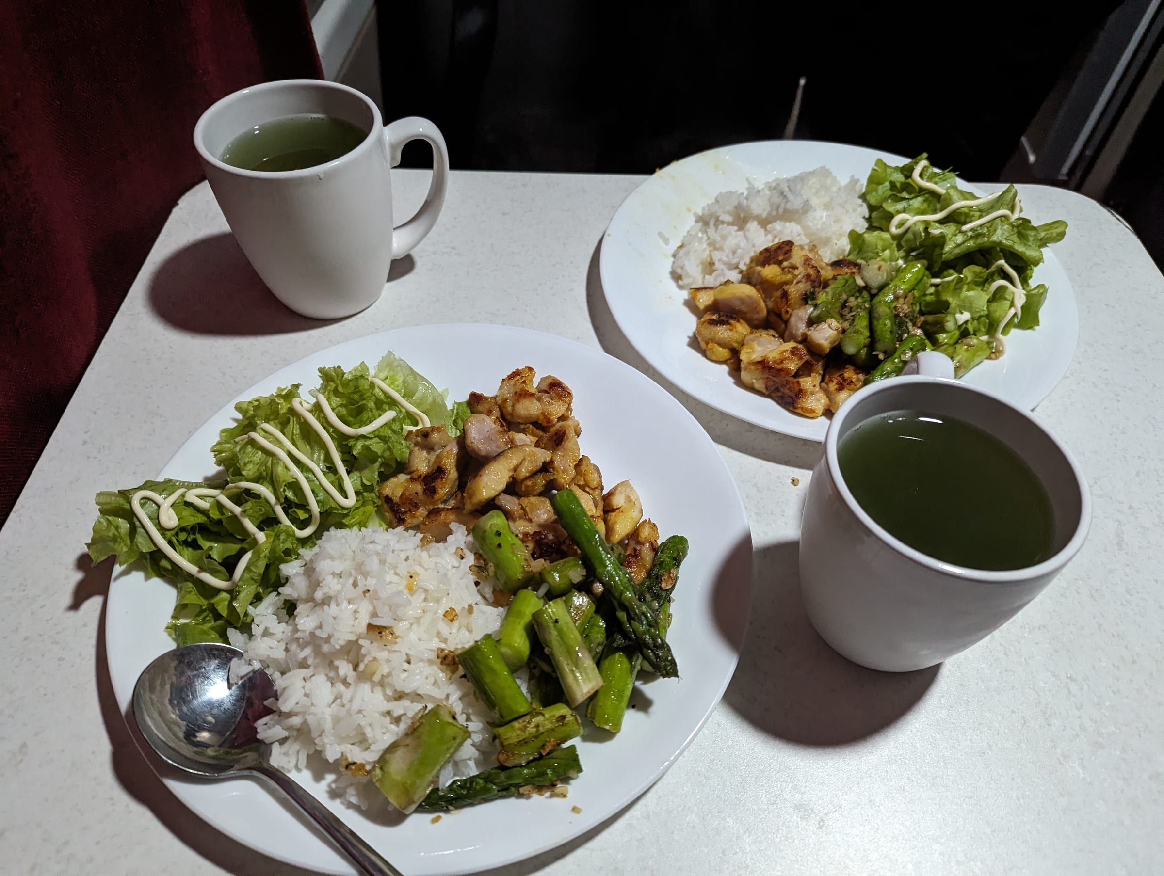 Two plates of stir-fried chicken, rice, and asparagus with mugs of tea, set up inside the van at night