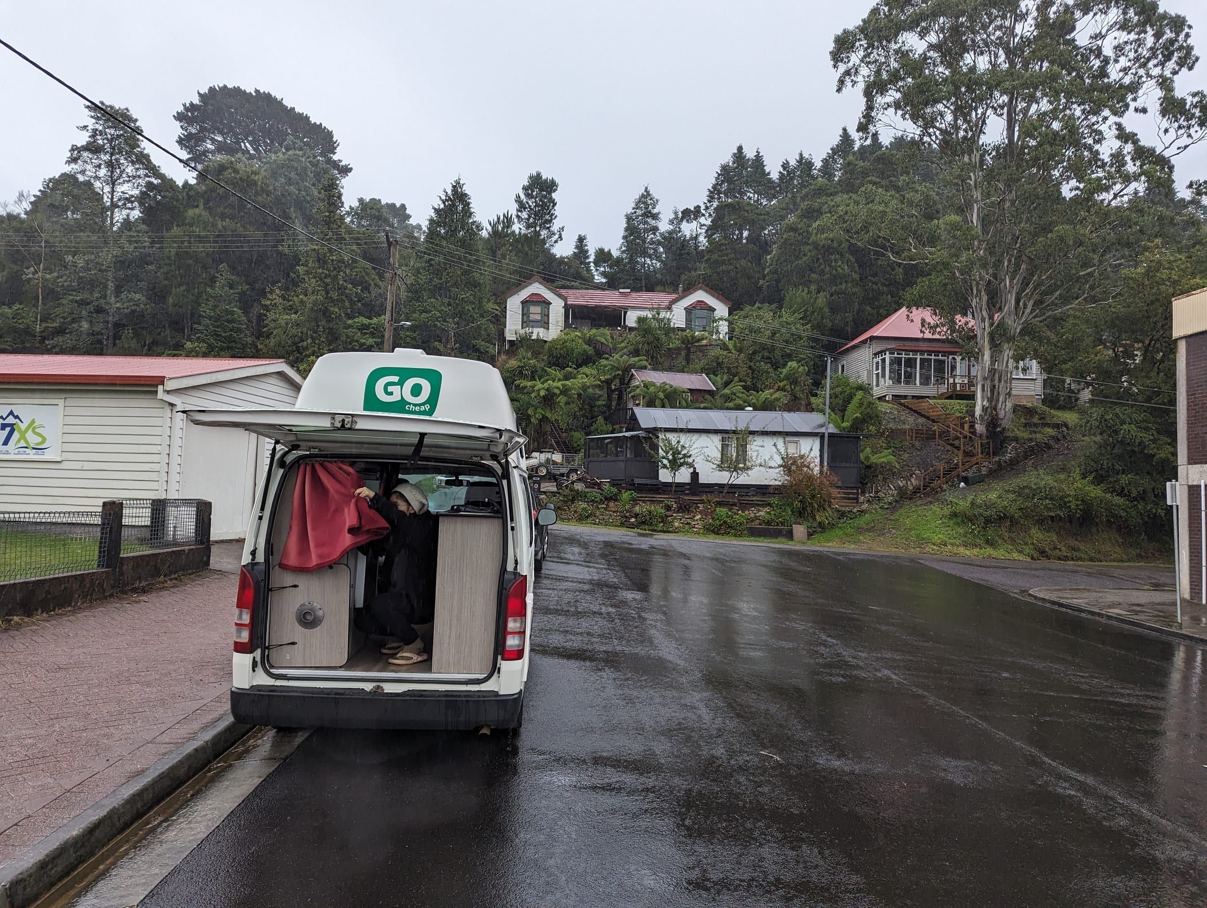The "GO Cheap" rental van parked on a quiet wet street, back doors open, rain falling